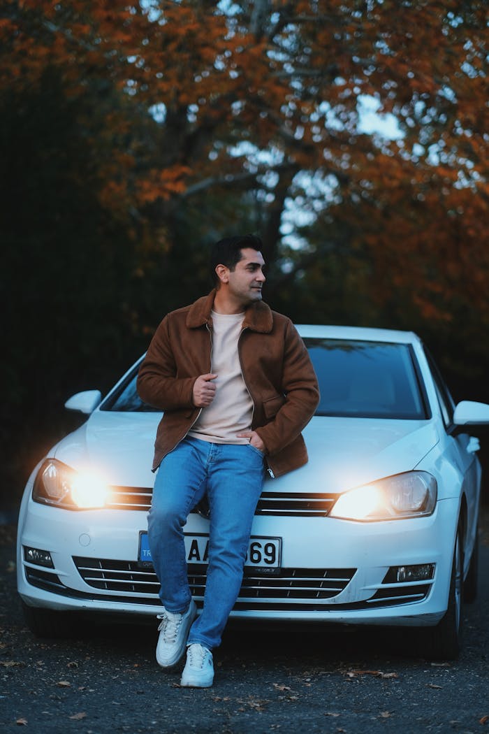 A man in a jacket leans against a car with autumn leaves in the background, captured outdoors in Türkiye.